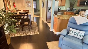 Un salon moderne avec un fauteuil bleu, un coin repas avec des chaises en bois, une cuisine équipée d’un réfrigérateur et un couloir menant à une autre pièce.