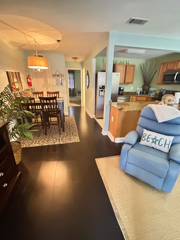 A modern living room with a blue armchair, a dining area with wooden chairs, a kitchen with a refrigerator, and a hallway leading to another room.