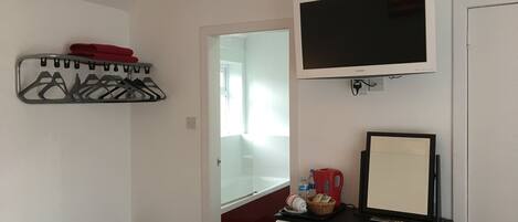 A compact room with a bed, a TV, a mirror, a shelf with hangers, a small table, and a chair.