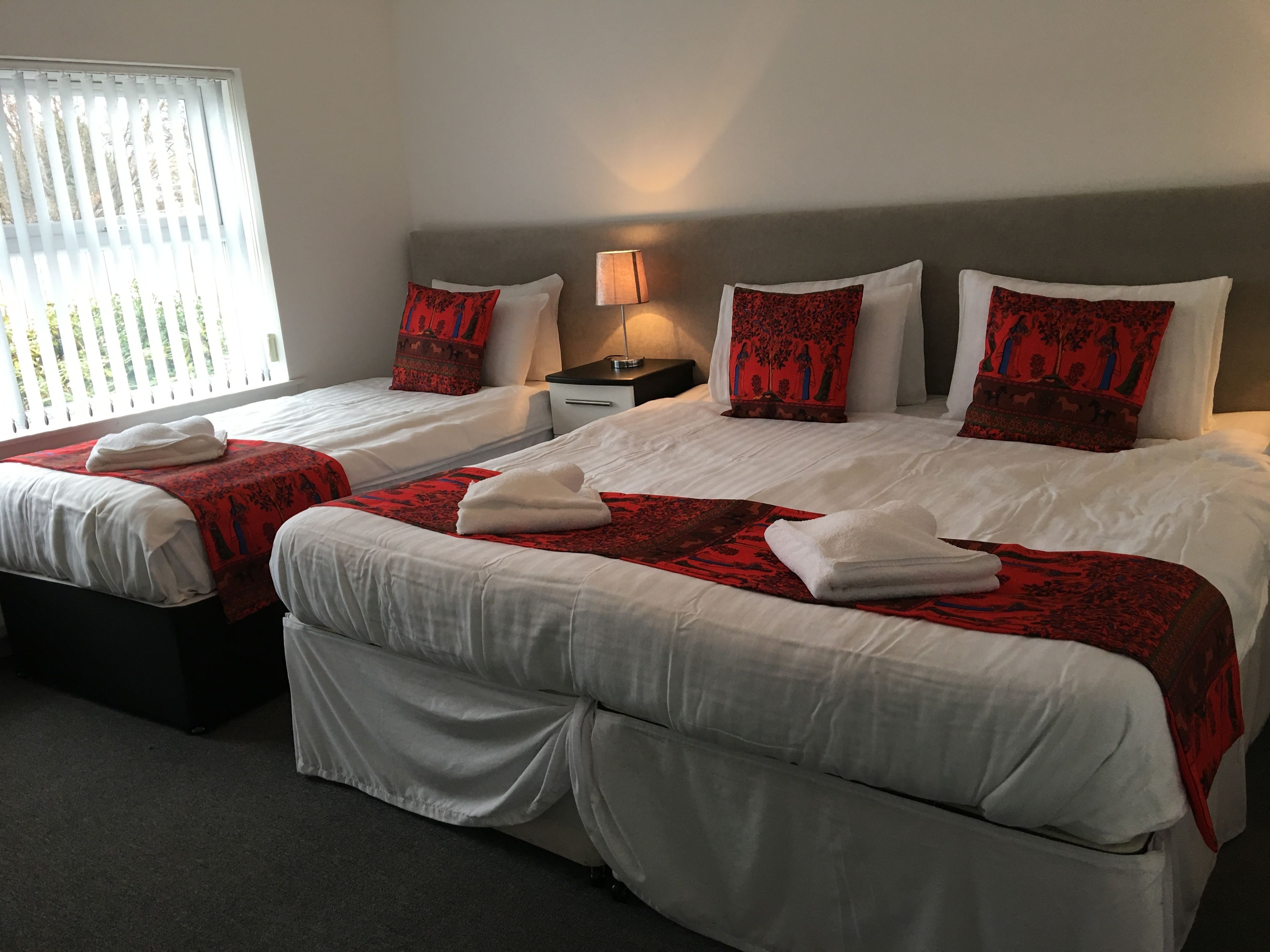 Habitación de hotel con dos camas, cada una con sábanas blancas y almohadones decorativos rojos. Entre las camas hay una mesita de noche con una lámpara.