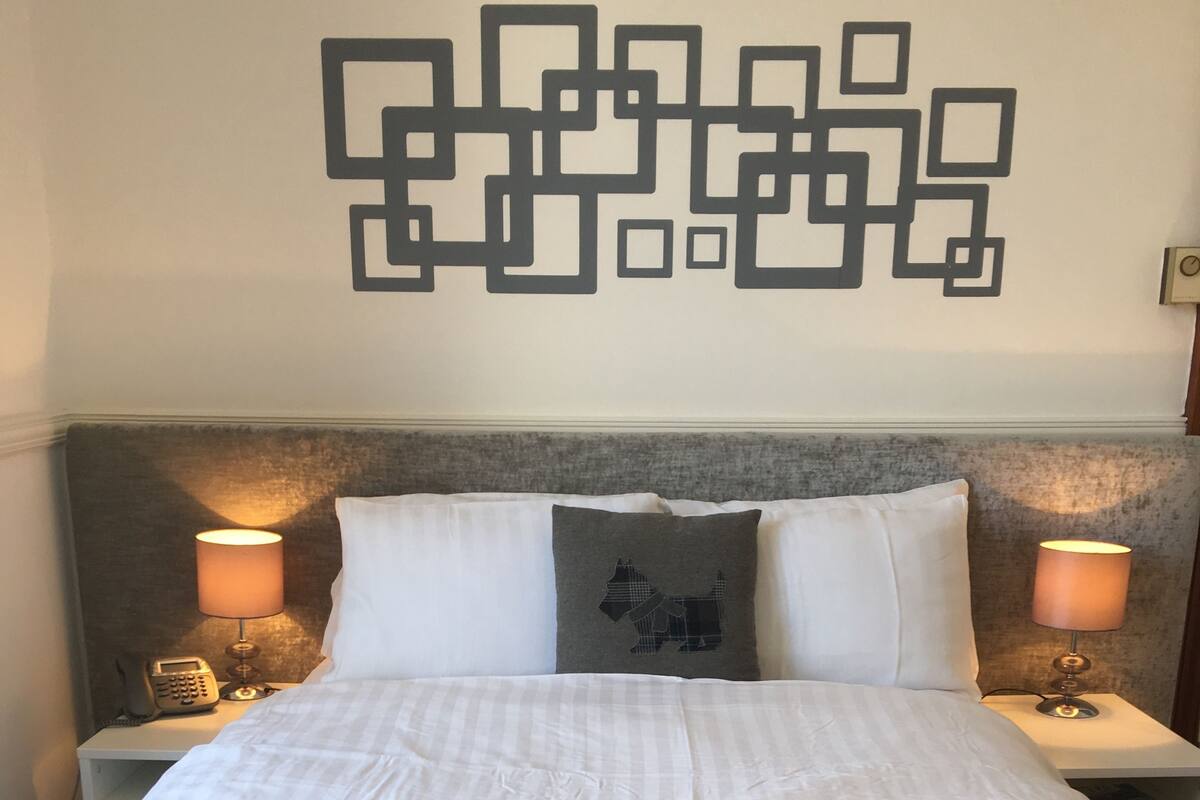 Un letto rifatto con lenzuola bianche, una coperta a quadretti grigia e nera, e un cuscino con un motivo a cavallo. Due abat-jour con paralumi, un telefono sul comodino e un quadro appeso sopra il letto.