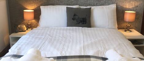 A neatly made bed with white linens, a gray and black checkered blanket, and a pillow with a horse design. There are two bedside lamps with lampshades, a telephone on the nightstand, and a wall art piece above the bed.