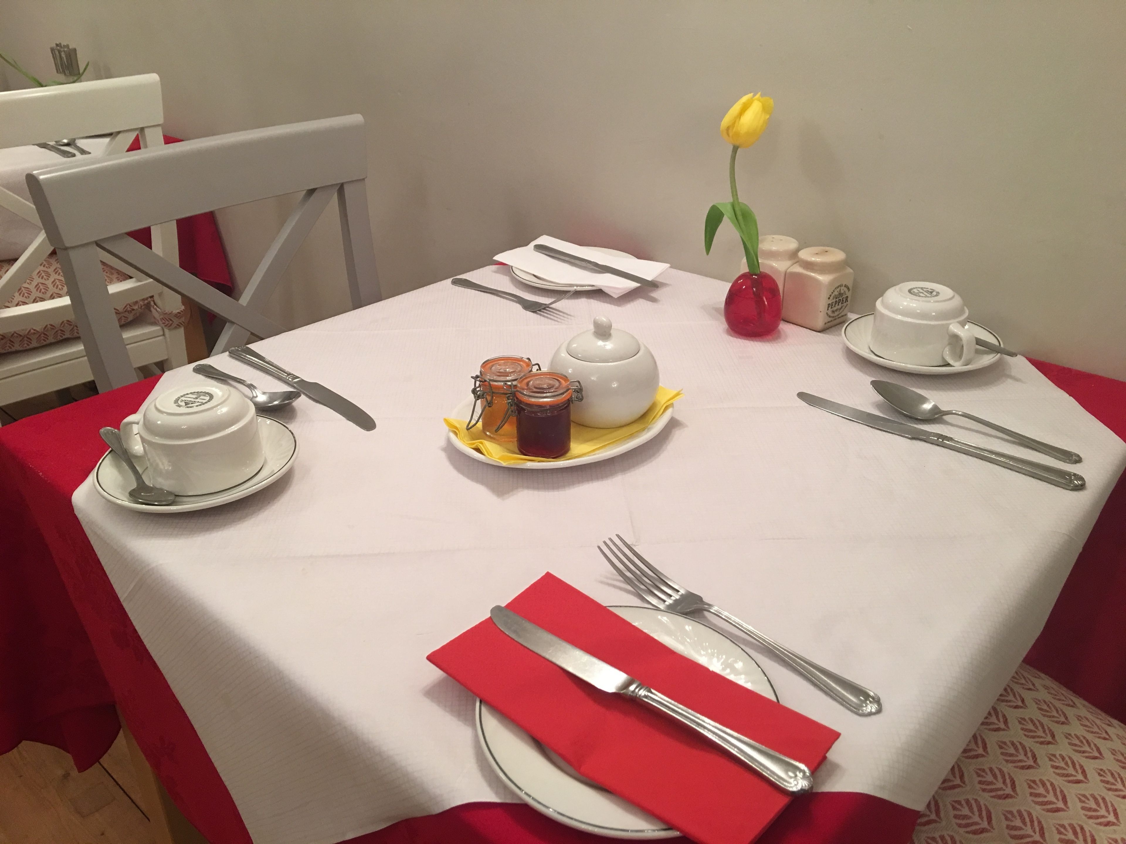 A neatly set dining table with a white tablecloth, plates, cutlery, a red napkin, a yellow tulip, and a white teapot.