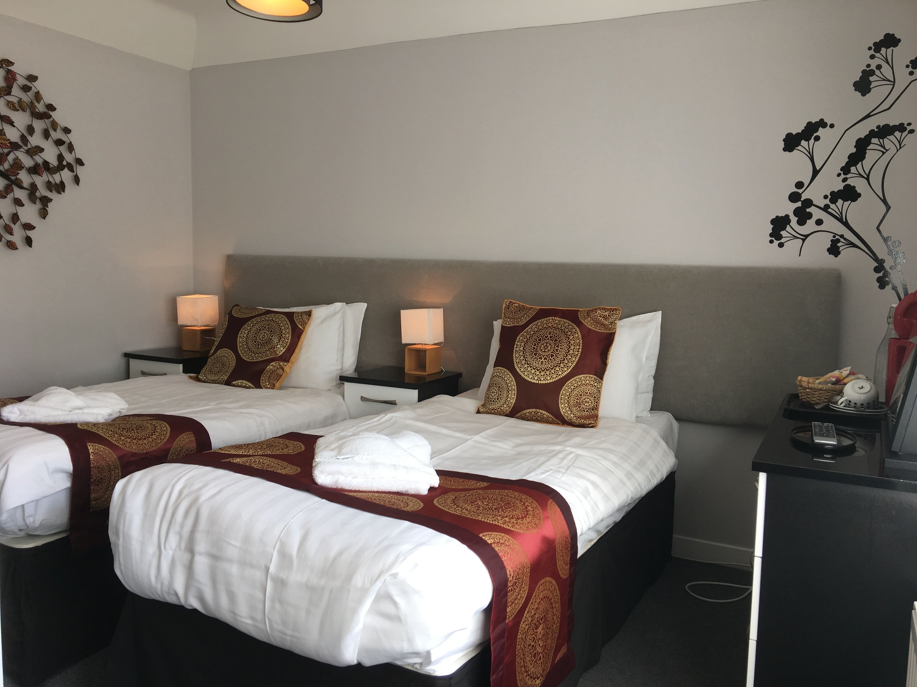 Habitación de hotel con dos camas, cada una con ropa blanca y almohadas decorativas. Hay mesitas de noche con lámparas y una pequeña mesa con una cesta y una tetera.