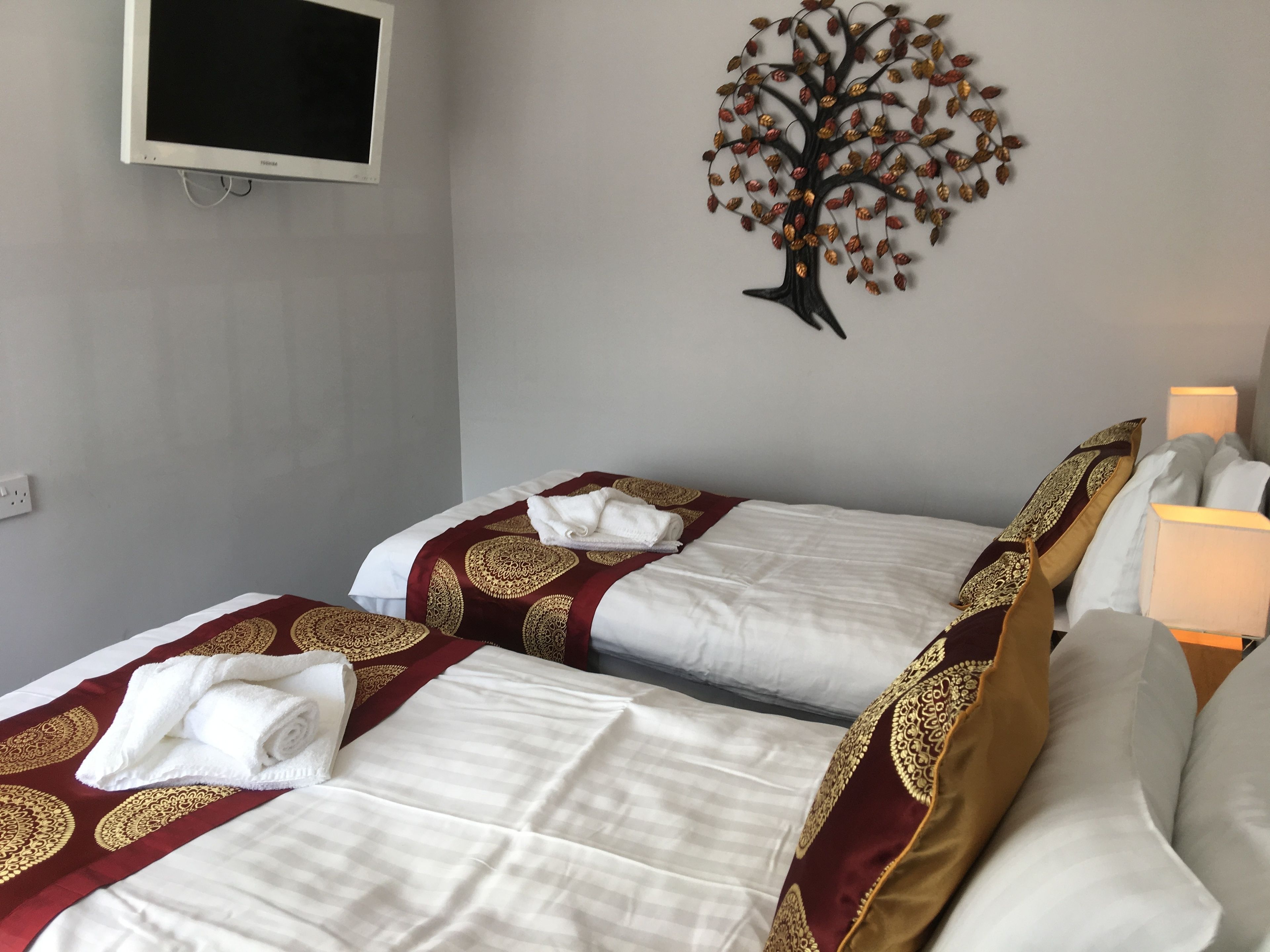 A hotel room with two beds, each with a white and maroon bedspread, and a decorative tree wall art.