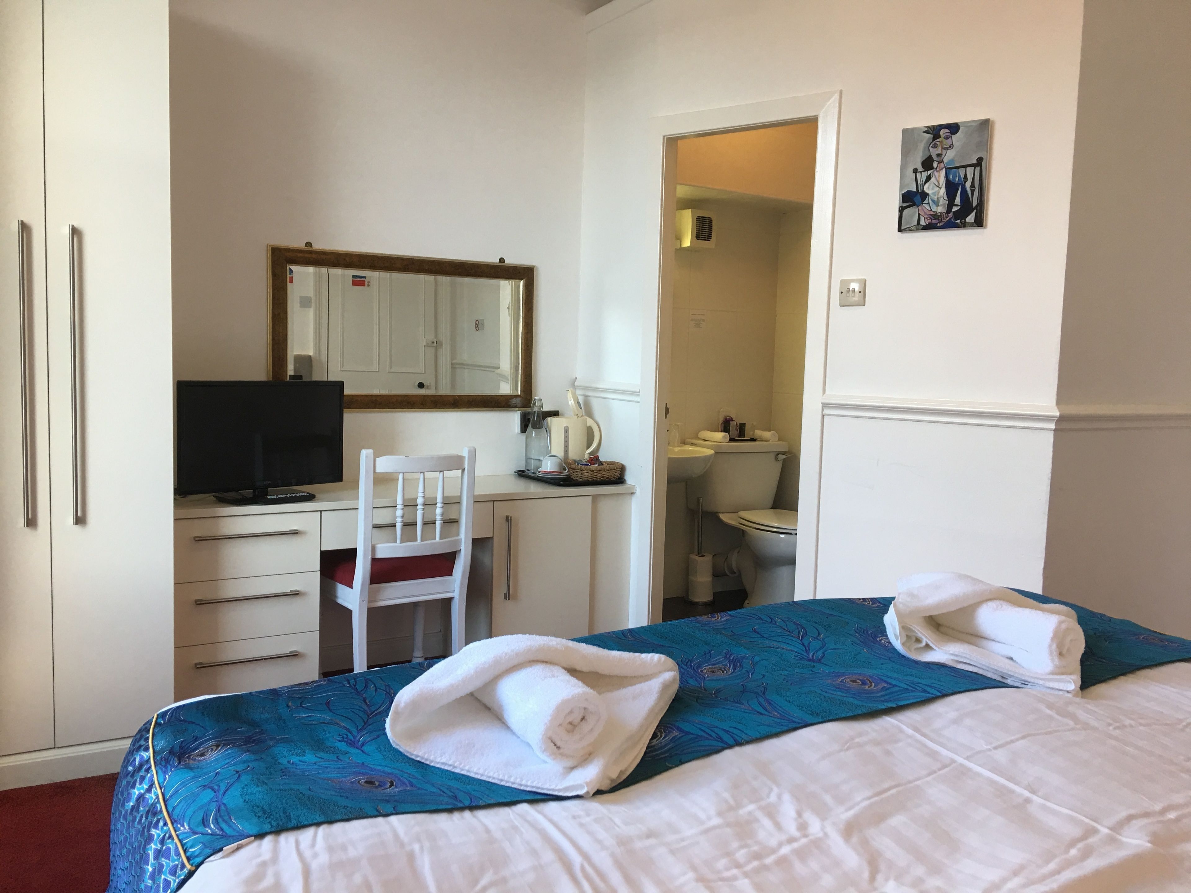 A hotel room with a bed, a desk with a computer, a chair, a bathroom, and a painting on the wall.