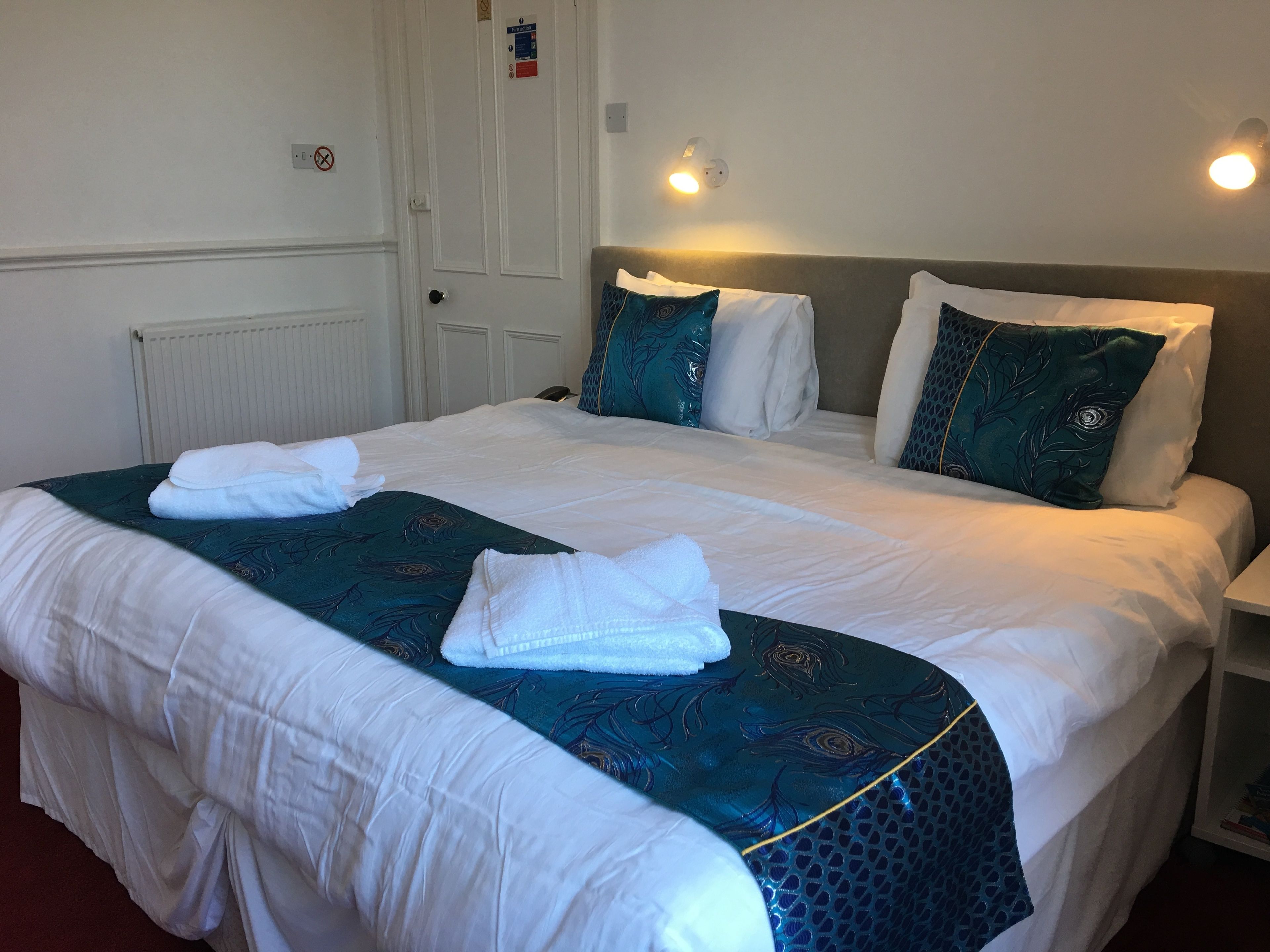 A neatly made bed with white linens and a teal-patterned bedspread, a bedside table with a lamp, and a door with a sign.