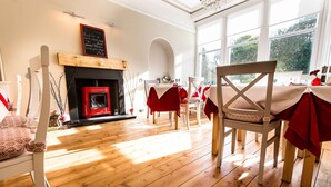 A dining area with wooden flooring, white chairs, a red tablecloth-covered table, and a fireplace with a red mantle. - Comrie Hotel (Crieff)