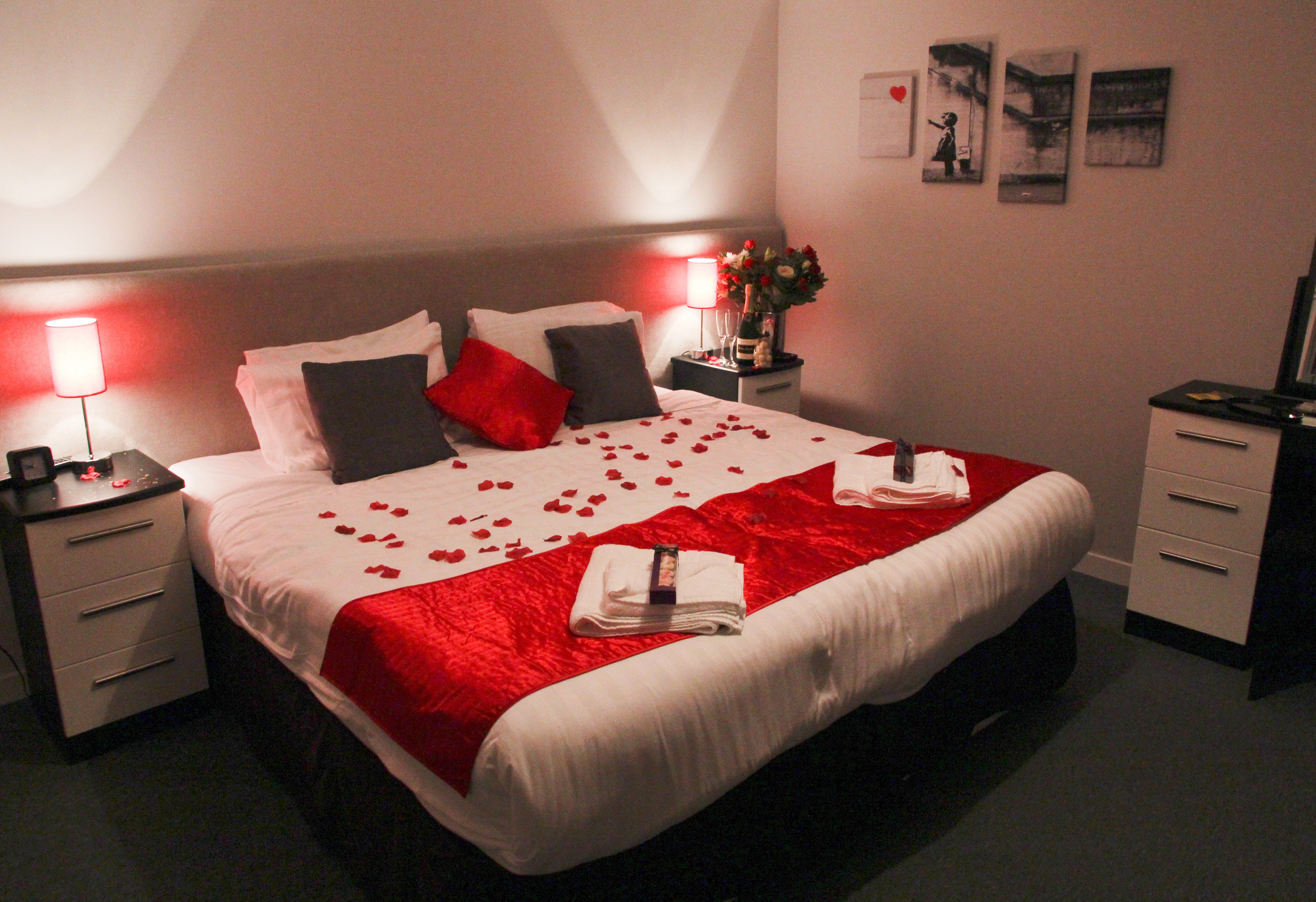 A neatly made bed with rose petals, a red blanket, and a bouquet of flowers on the nightstand.