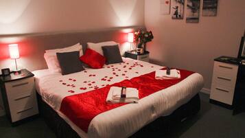 Una cama bien hecha con pétalos de rosa, una manta roja y un ramo de flores sobre la mesita de noche.