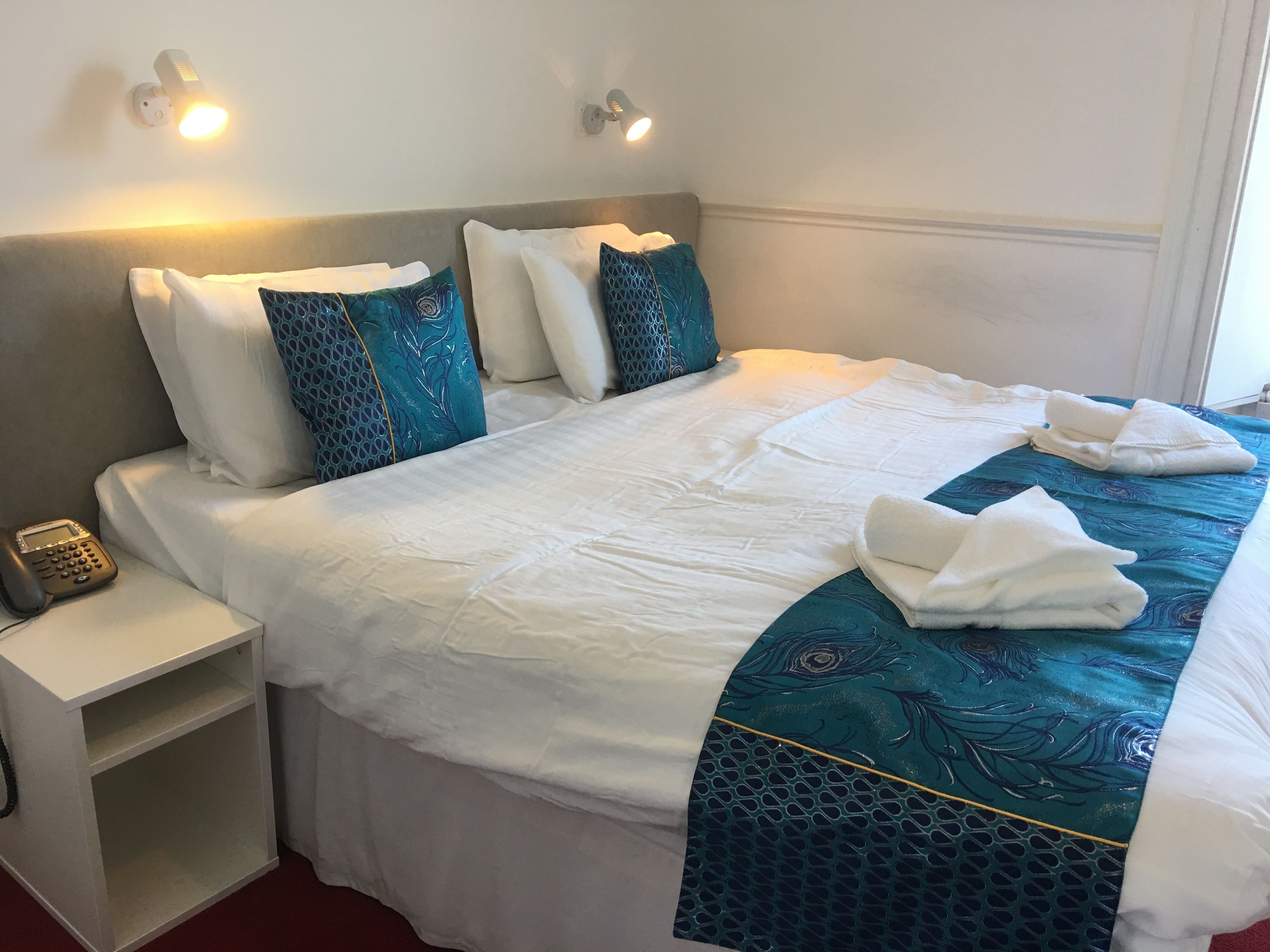 A neatly made bed with white and teal bedding, two wall-mounted lights, a bedside phone, and a small white nightstand.