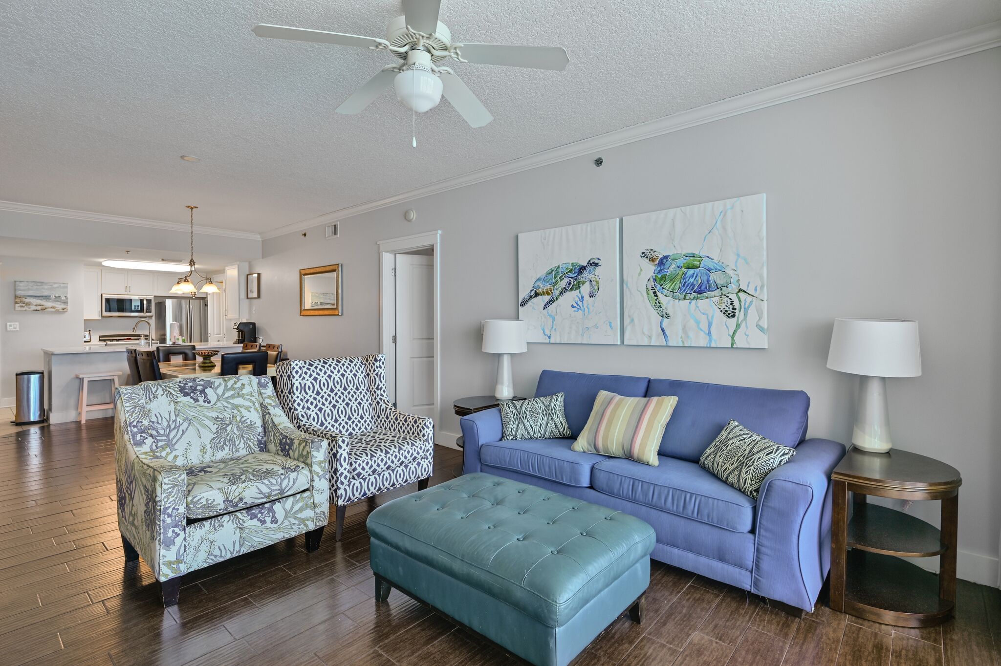 A living room with a blue sofa, patterned armchair, and ottoman. There is a ceiling fan, artwork on the wall, and a kitchen in the background.