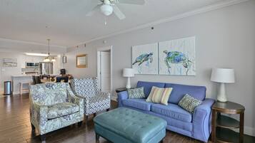 A living room with a blue sofa, patterned armchair, and ottoman. There is a ceiling fan, artwork on the wall, and a kitchen in the background.