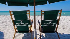 Twee groene strandstoelen met het Waterscape-logo onder een groene parasol op een zandstrand.