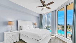 Quarto com cama grande, mesa de cabeceira, ventoinha no teto e vista para uma piscina.