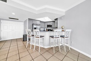 A kitchen with a central island, white bar stools, and a tiled floor.