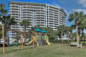 A playground with slides and swings, a bench, and palm trees in front of a large hotel building.