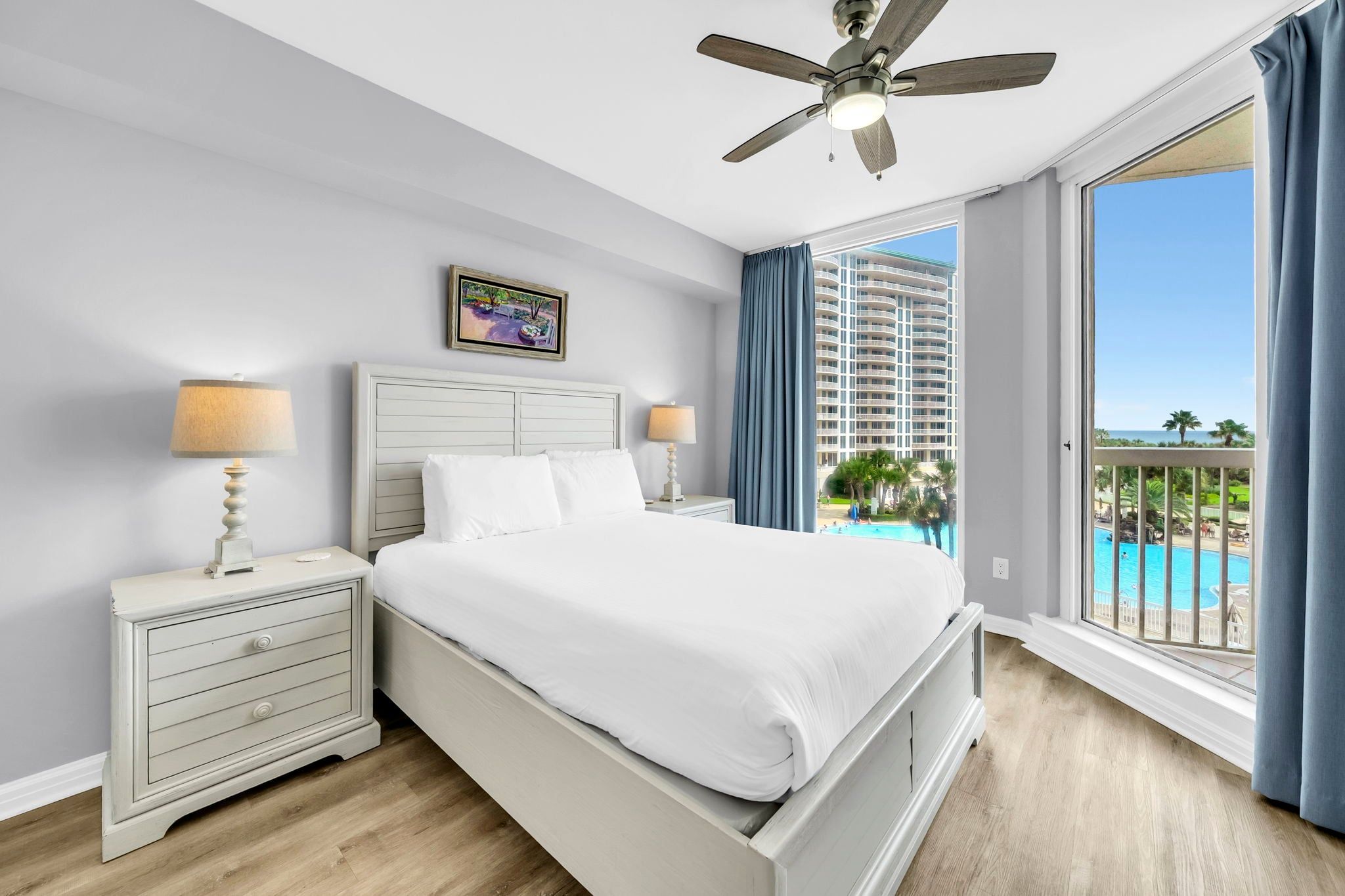 Ein Hotelzimmer mit einem Bett, zwei Nachttischen mit Lampen, einem Deckenventilator, einem großen Fenster mit Blick auf einen Pool und Gebäude sowie einem Gemälde an der Wand.