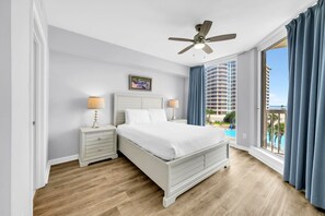 Une chambre d’hôtel avec un lit, deux tables de chevet, un ventilateur de plafond, un tableau et une vue sur un immeuble et une piscine.