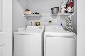 A laundry area with two front-loading washing machines, a shelf with cleaning supplies, and a utility wall with a water heater and electrical outlets.