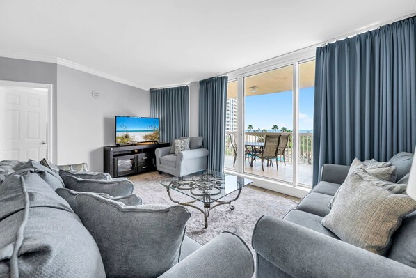 Ett modernt vardagsrum med en stor platt-TV, grå soffor, ett soffbord i glas och en balkong med utsikt över stranden.