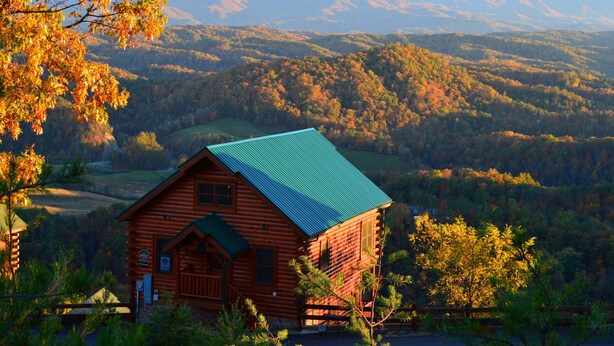 A log cabin with a green roof, surrounded by autumn foliage and mountains in the background. - 'Mountainscape'-Million Dollar Views In Legacy Mtn. 10% off 7 night rental! (Pigeon Forge)