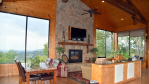 A spacious living room with a stone fireplace, large windows, wooden ceiling, and a television mounted above it. - 'Mountainscape'-Million Dollar Views In Legacy Mtn. 10% off 7 night rental! (Pigeon Forge)