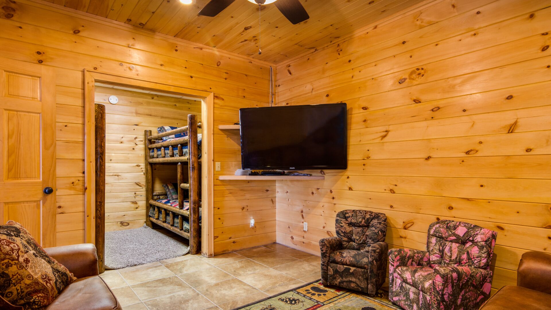 A cozy cabin interior with a TV mounted on the wall, a wooden door, a bookshelf, and two patterned armchairs.