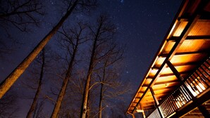 Eine Holzhütte mit Balkon, umgeben von hohen Bäumen unter einem sternenübersäten Nachthimmel.