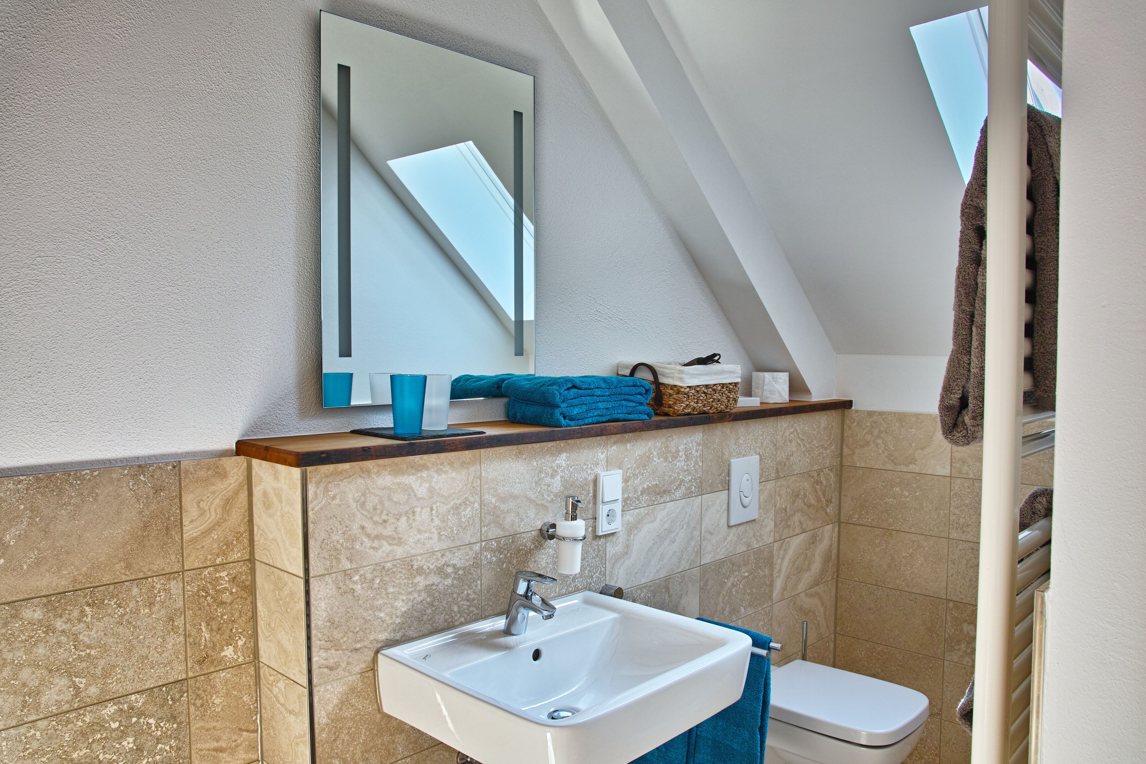 A modern bathroom with a white sink, a mirror, and a toilet. There is a towel rack and a shelf with toiletries.