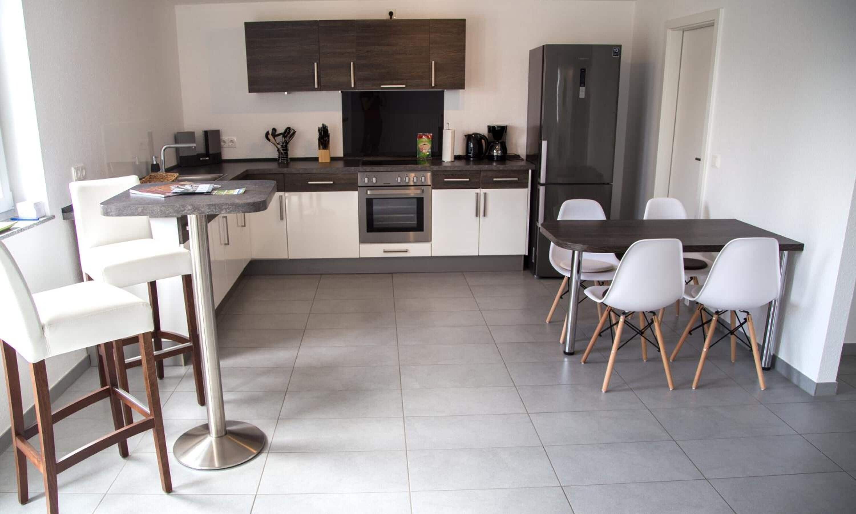 A modern kitchen with white and dark wood cabinets, a stainless steel oven, a refrigerator, and a dining area with white chairs and a dark table.