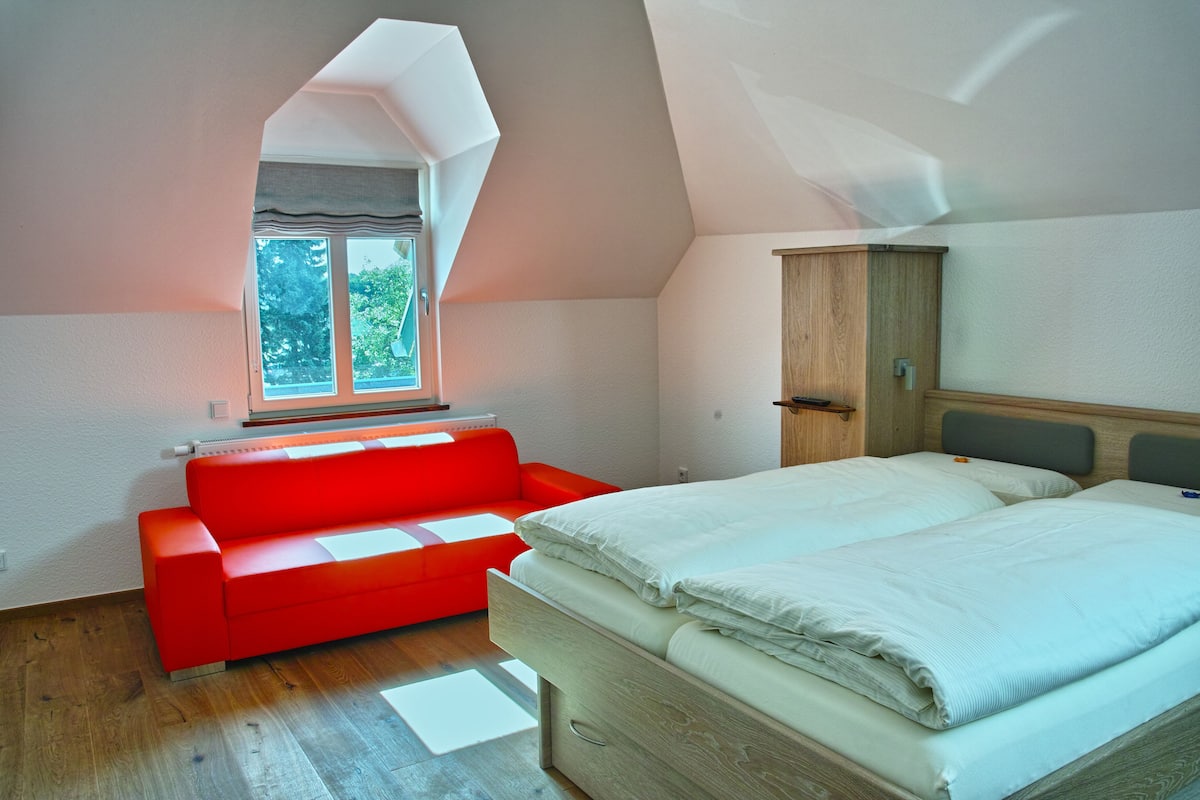A bedroom with a loft bed, a red sofa, a wooden wardrobe, and a window with a view of trees.