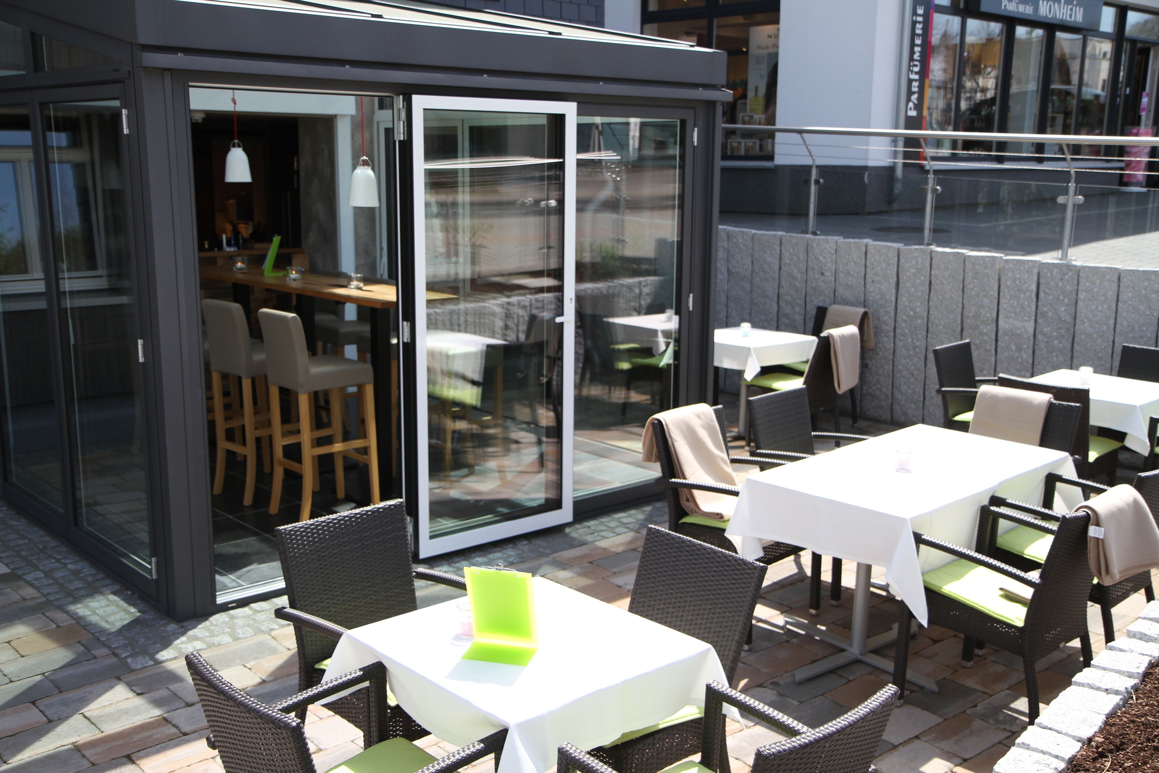 Un espace de restauration en plein air doté de coins repas et de tables protégés par des parois de verre, avec des chaises en osier et des nappes blanches.