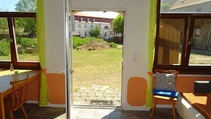 A room with a wooden table and chairs, a window with curtains, and a view of a building and garden outside. - Elbschloss Kehnert (Tangerhütte)