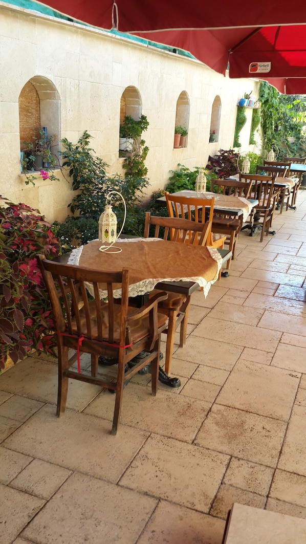 A row of wooden tables and chairs under a red awning, with potted plants and a tiled floor. - Elif Hatun Konagi (Tarsus)