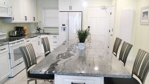 A modern kitchen with white cabinets, a granite countertop, and a central island with chairs. - #102 First Floor, Direct Ocean Front, 2 King Beds, 1 Queen Murphy, Dog Free 20# (Daytona Beach)