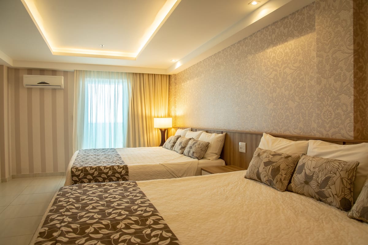 Ein Hotelzimmer mit zwei Betten, einer Deckenleuchte, einem Fenster mit Vorhängen und einer Wand-Klimaanlage.