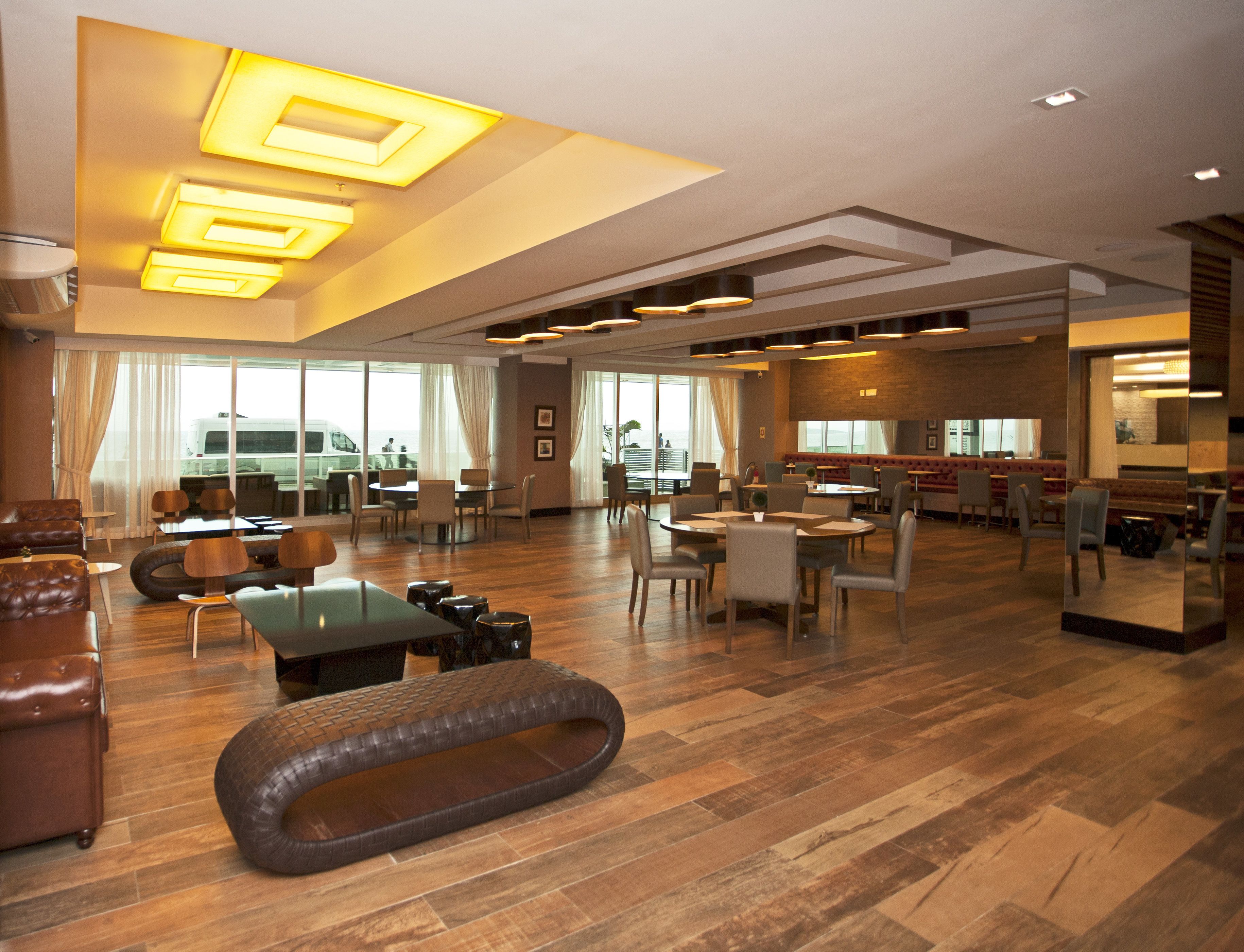 A spacious hotel lobby with a central seating area, multiple dining tables, and a large window offering a view of the outside.