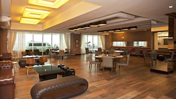 A spacious hotel lobby with a central seating area, multiple dining tables, and a large window offering a view of the outside.