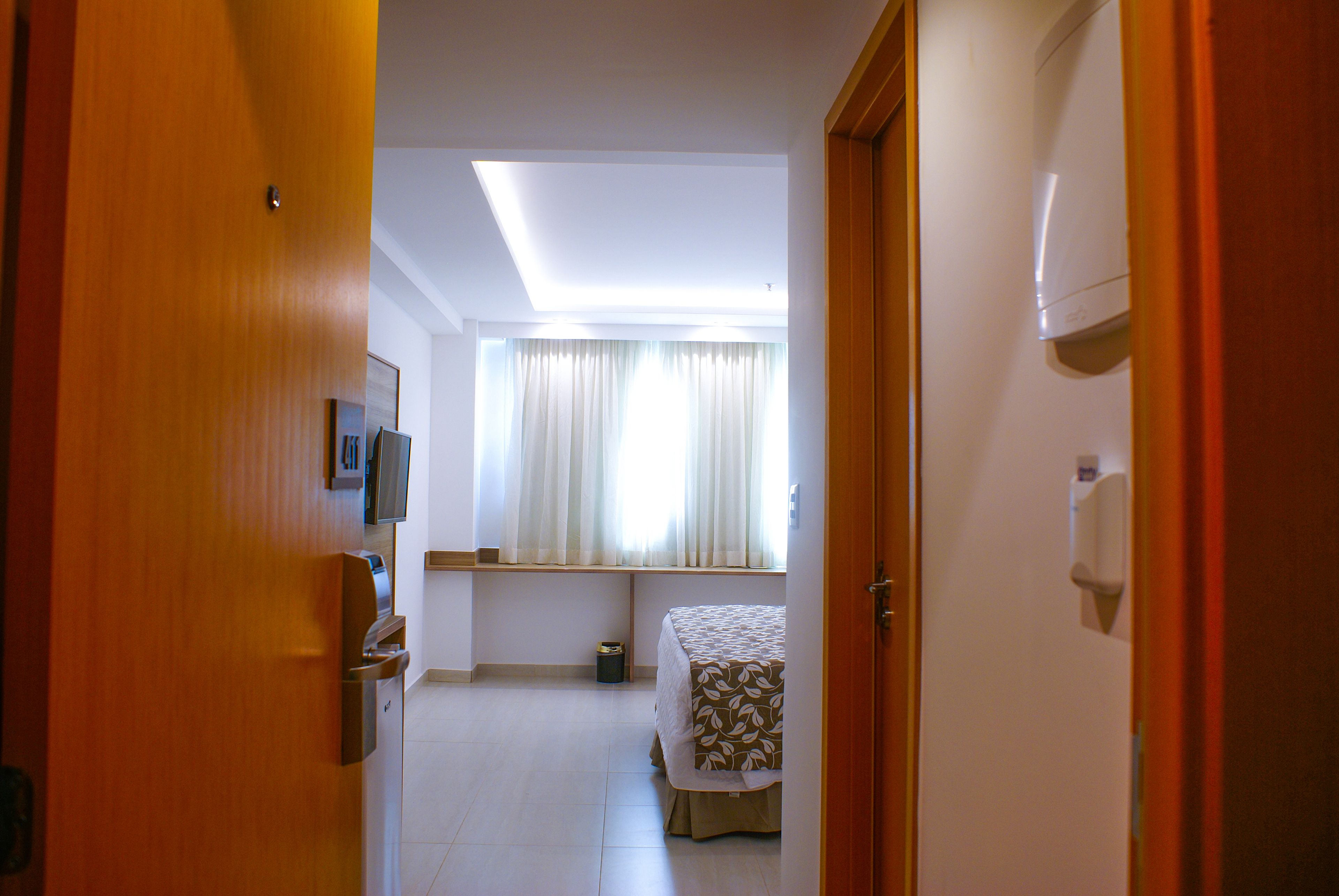 A hotel room with a bed, a small table, a window with curtains, a ceiling light, and a door.