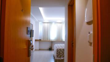 A hotel room with a bed, a small table, a window with curtains, a ceiling light, and a door.