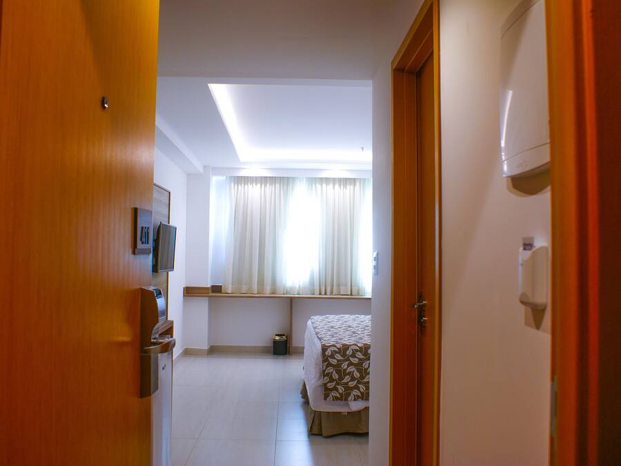 A hotel room with a bed, a small table, a window with curtains, a ceiling light, and a door.
