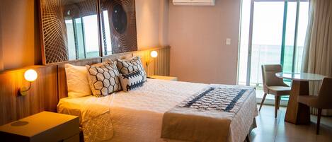 A hotel room with a large bed, a desk, and a chair. There is a window with curtains and an air conditioning unit on the wall.