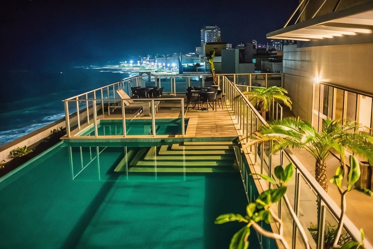 A rooftop pool with a city view, outdoor seating, and palm plants.