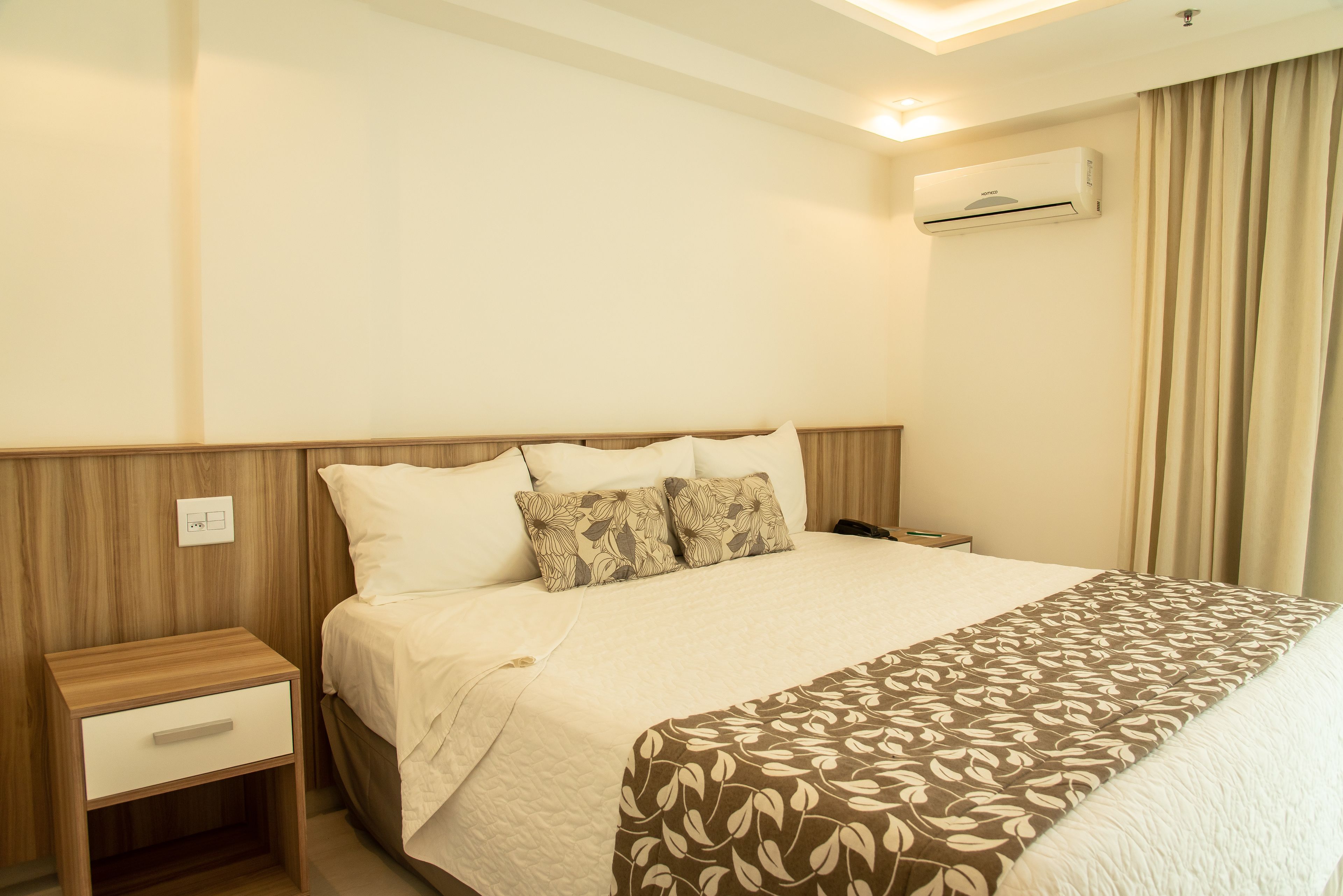 A neatly made bed with a patterned bedspread, a wooden nightstand with a drawer, and an air conditioning unit on the wall.