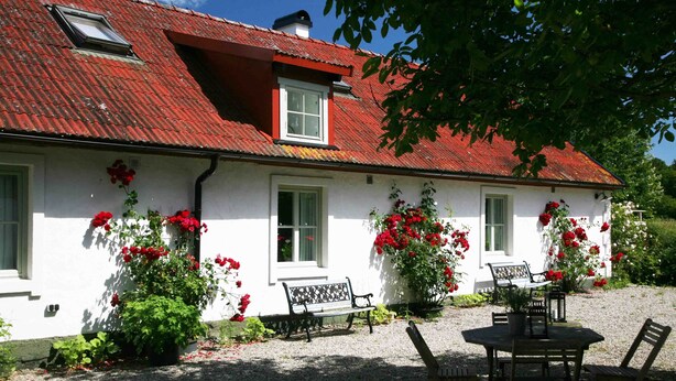 A white house with a red-tiled roof, a bench, and a table with chairs in the front yard. - Stenrosgården logi (Kivik)