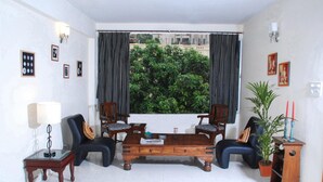 Un salon avec une table basse en bois, deux fauteuils, une table d’appoint, une lampe et une vue sur la verdure à travers les rideaux.