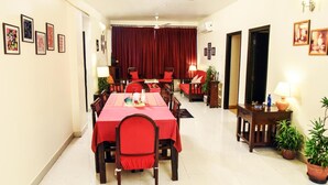 Un coin repas avec une longue table, des chaises rouges et une nappe rouge. Les murs sont ornés de tableaux encadrés et un ventilateur de plafond est installé au centre.