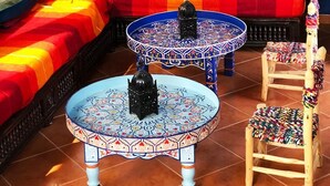 A room with a blue wall, two windows, a sofa with colorful cushions, a patterned table, and a chair.