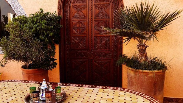 A traditional Moroccan door with intricate carvings, surrounded by potted plants and a tiled table with a tray of tea and glasses.